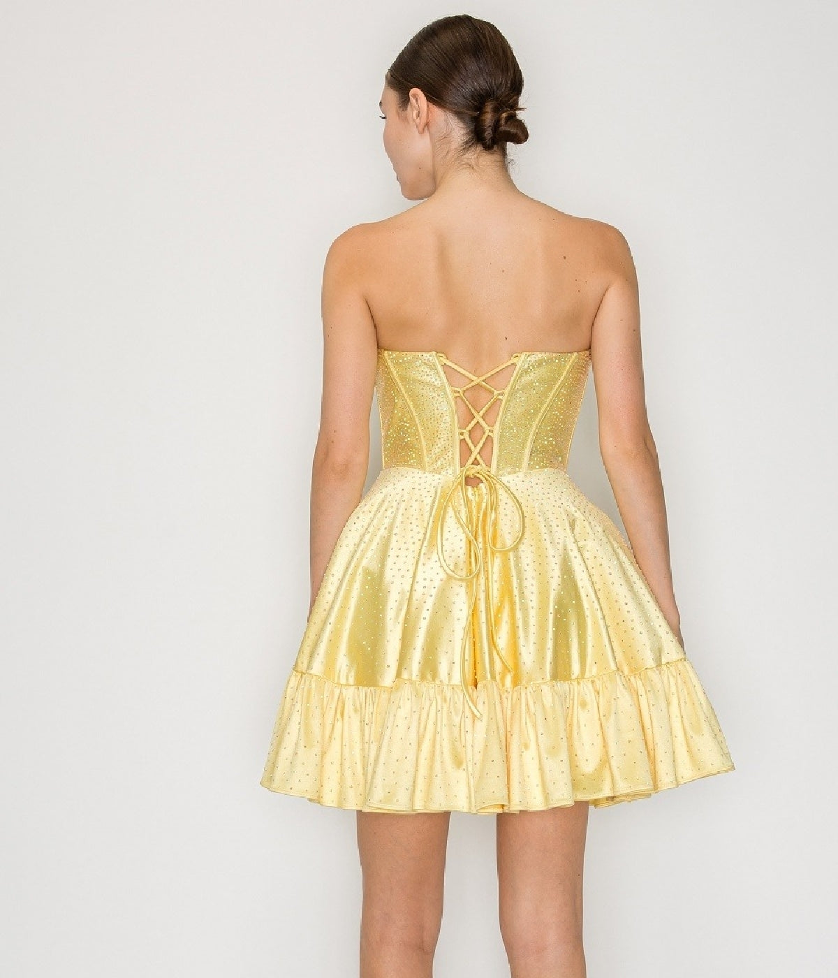 A woman stands facing away, wearing the Poly USA 9942 short homecoming dress in yellow, featuring a strapless design, lace-up back, and ruffled hem.